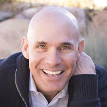 A close-up portrait of a smiling man wearing a dark sweater over a collared shirt. He is outdoors with a blurred natural background of rocks and grass.