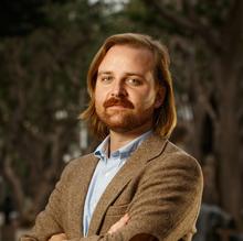 Portrait of a person with shoulder-length red hair and a mustache, wearing a brown blazer over a light blue shirt. They have a confident expression with arms crossed, standing outdoors with blurred trees in the background.